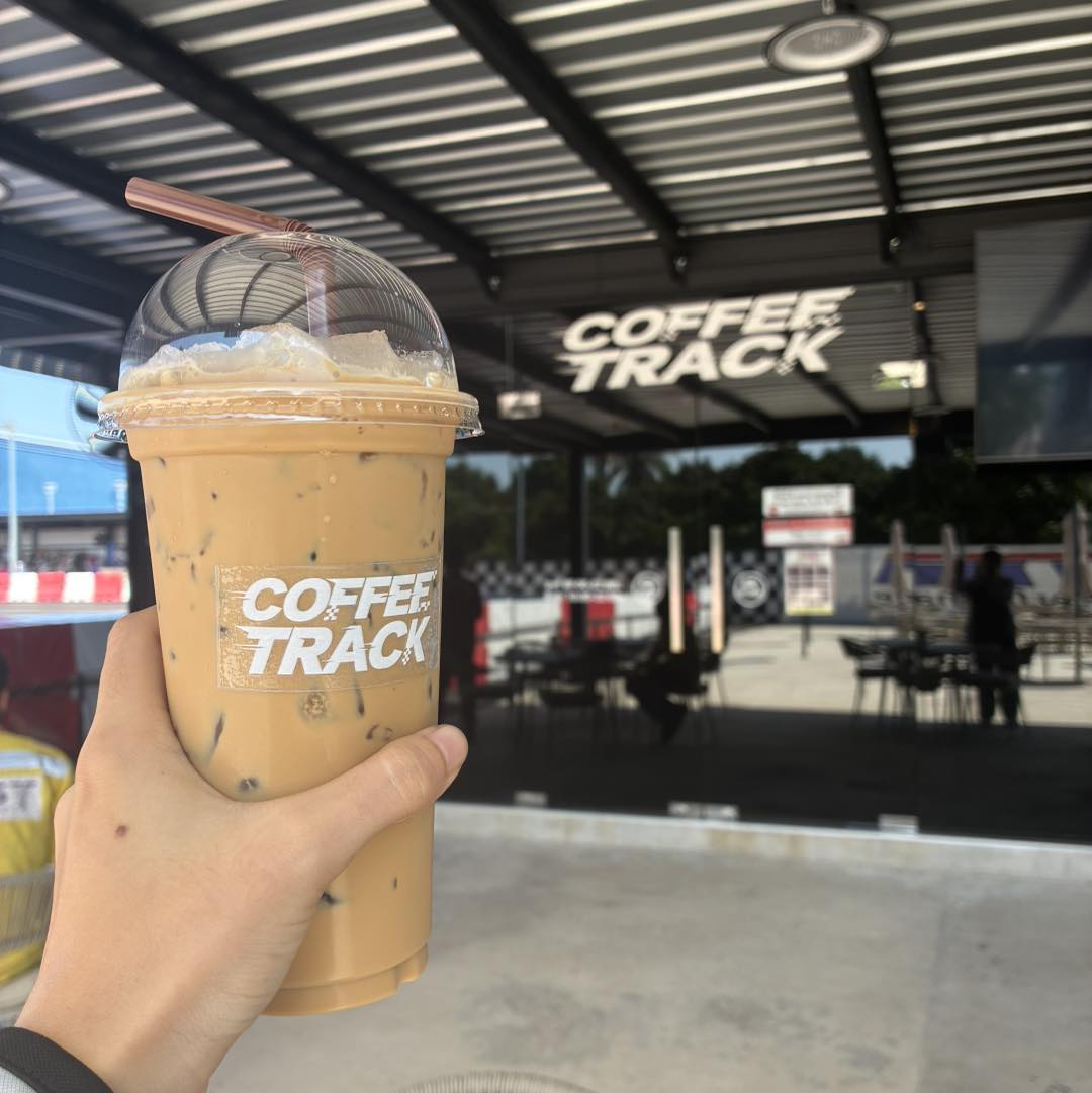 กาแฟเย็น Coffee Track แก้วแบรนด์ กับวิวคาเฟ่และสนามแข่งด้านหลัง / Coffee Track iced coffee in a branded cup with the cafe and circuit behind