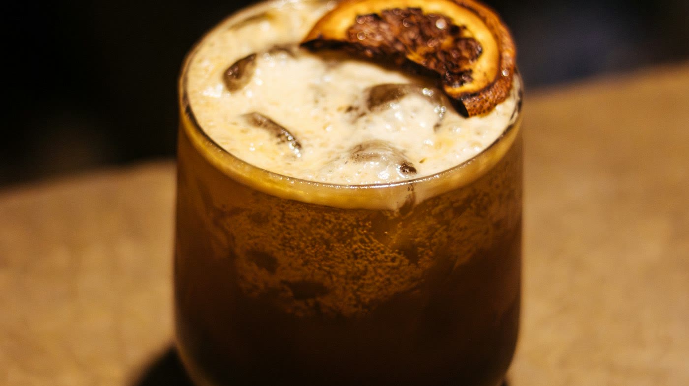 กาแฟดำเย็นในแก้วใส มีครีมด้านบนและส้มอบแห้งตกแต่ง (ภาพประกอบ) / Iced black coffee in a clear glass with crema and a dried orange slice (illustrative stock image)