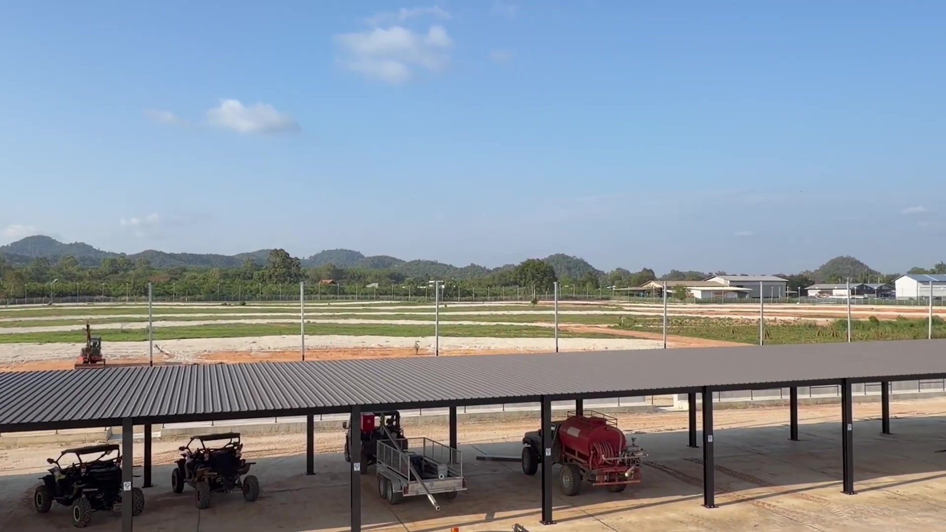 ภาพถ่ายจริงหน้างานก่อสร้าง EEST&nbsp;Circuit — โครงหลังคาพิทเหล็กและพื้นที่ปรับหน้าดินสำหรับสร้างสนาม พร้อมภูเขาเป็นฉากหลัง / Real on-site photo of the EEST&nbsp;Circuit build — steel pit-roof frame and graded earthworks with mountains behind